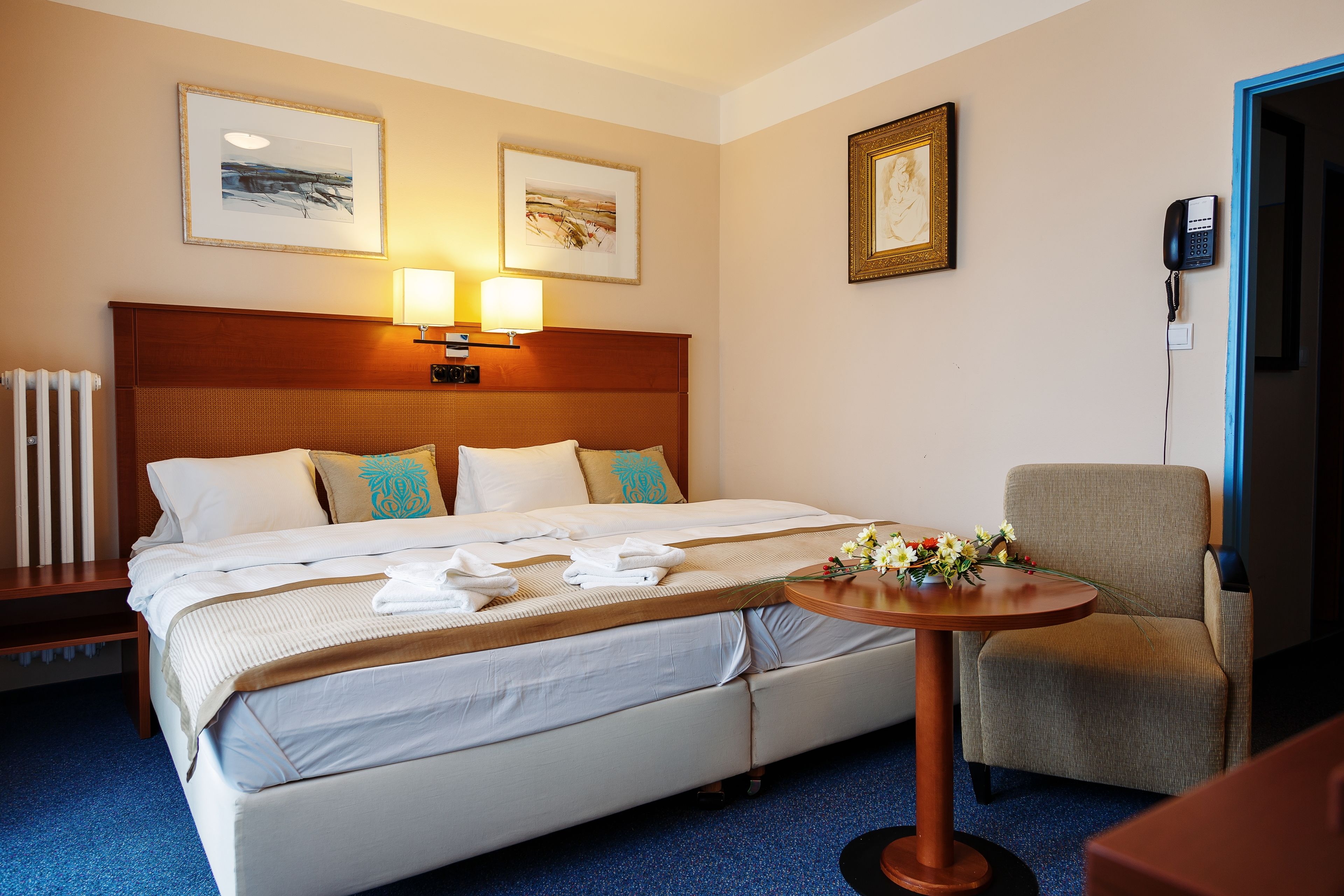 A hotel room with a bed, bedside lamps, a chair, a table with flowers, and artwork on the walls.