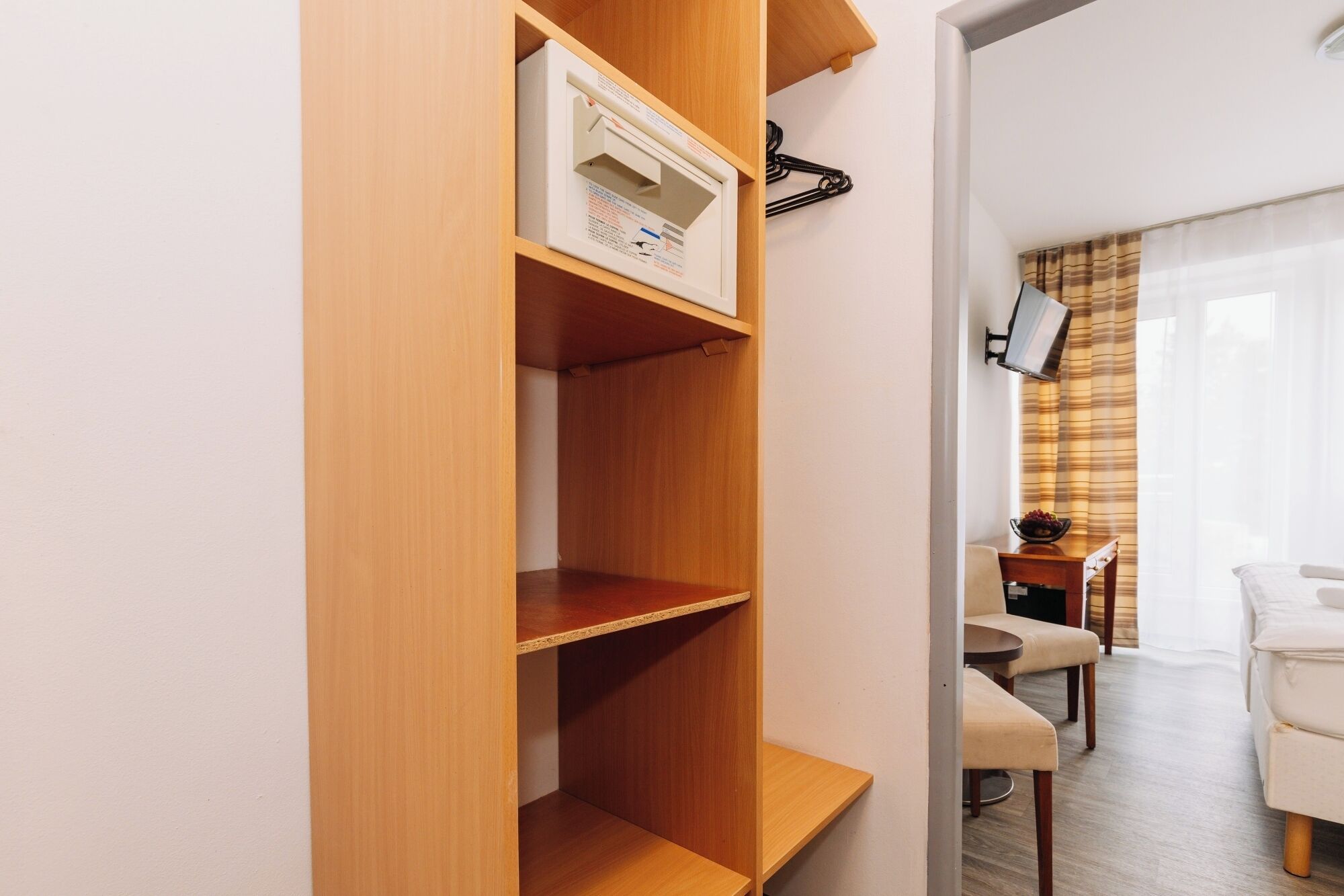 Een hotelkamer met een houten garderobe, een wandtelevisie, een eethoek met tafel en stoelen, en een raam met gordijnen.