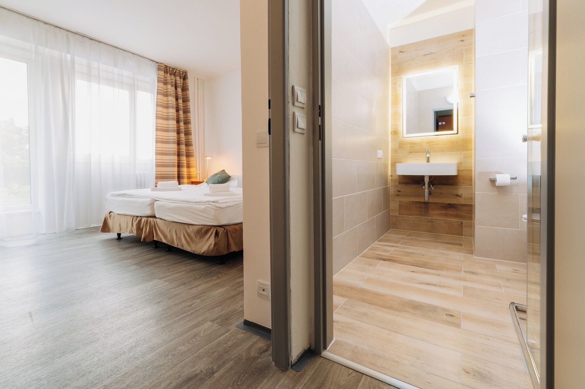 Une chambre d’hôtel avec un lit, une salle de bain comprenant un lavabo et un miroir, et un plancher en bois.