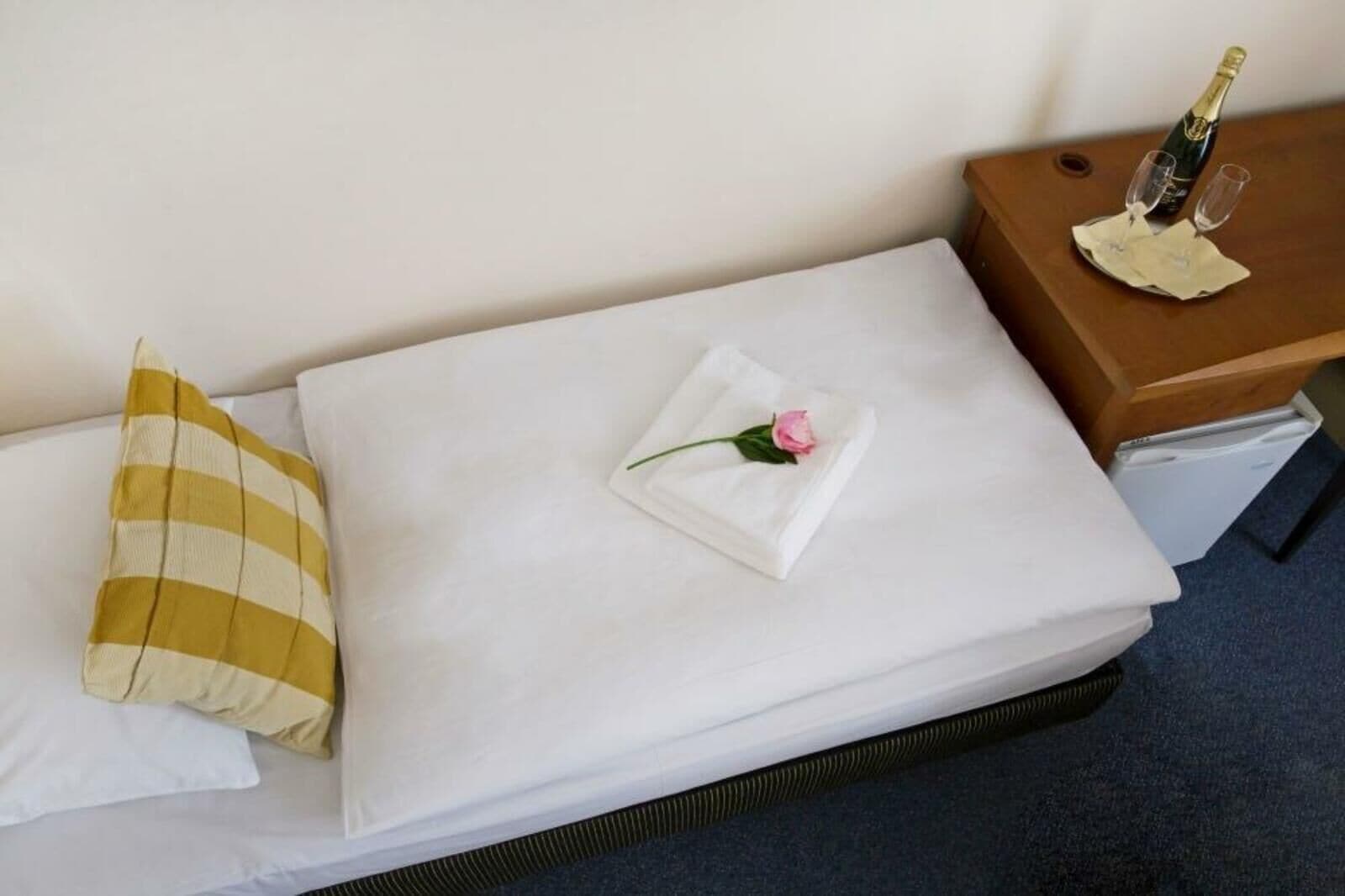 Un lit bien fait, avec un oreiller blanc, un oreiller à rayures jaunes et une serviette blanche pliée, ornée d’une rose rose. On y voit une table de chevet en bois sur laquelle sont posés une bouteille de champagne et deux verres.