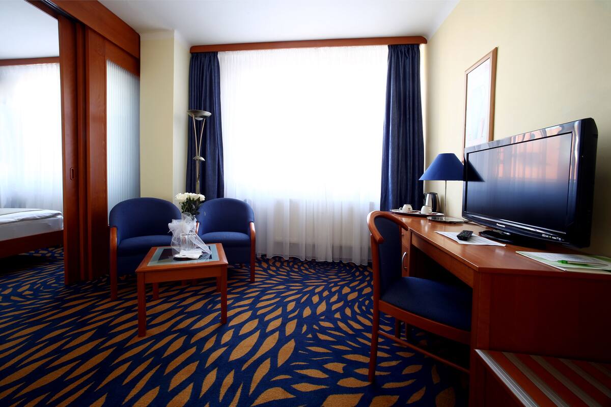 Ein Hotelzimmer mit einem Bett, zwei blauen Sesseln, einem kleinen Tisch mit Blumenvase, einem Schreibtisch mit Fernseher und einem großen Fenster mit Vorhängen.