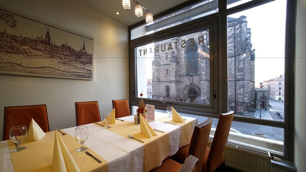A restaurant with a table set for four, a large window with a view of a historic building, and a cityscape painting on the wall. - Central Hotel Pilsen (Plzen)