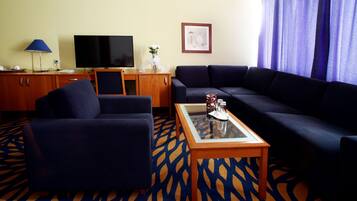 Habitación de hotel con un sofá azul oscuro, una mesa de centro de madera, un televisor sobre un mueble de madera y una alfombra estampada.