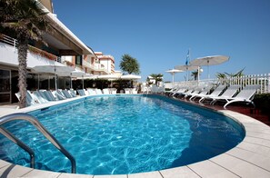 A swimming pool with lounge chairs and umbrellas, surrounded by a white fence and palm trees. - Hotel Rivamare (Jesolo)