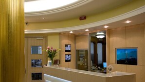 A reception area with a modern reception desk, a large pillar, a reception desk with a vase of flowers, and a wall-mounted television. - Hotel Rivamare (Jesolo)
