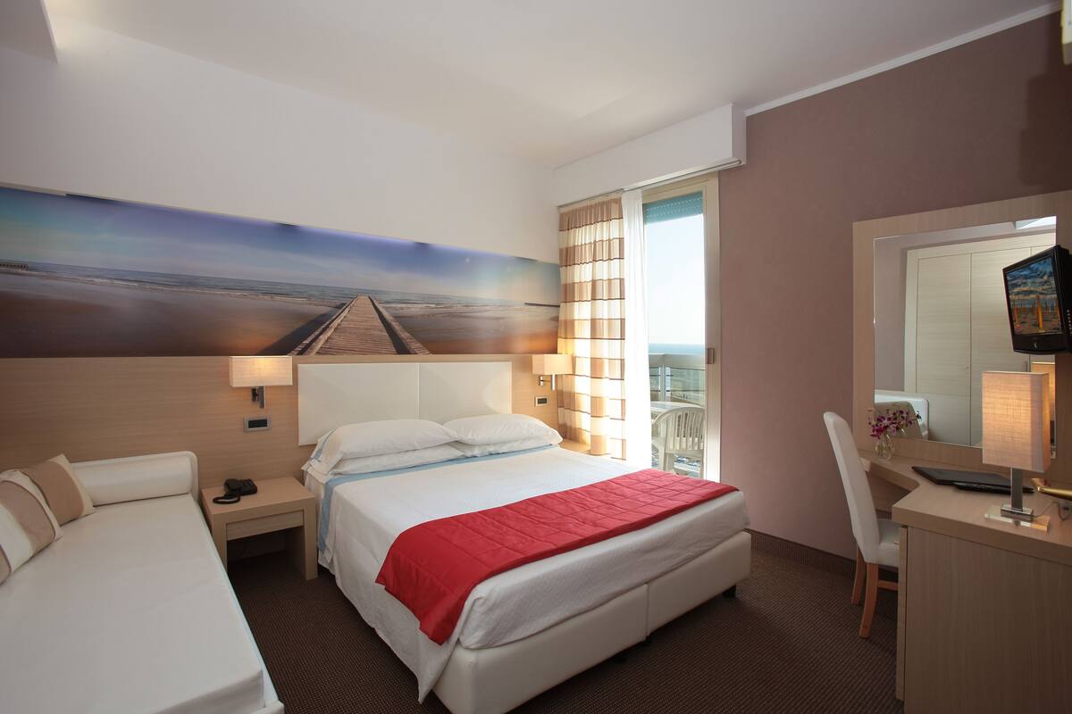 Une chambre d’hôtel avec un lit, un bureau, une chaise, une télévision et un tableau représentant une scène de plage.