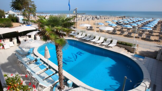 A pool area with lounge chairs and umbrellas, a palm tree, and a view of the beach and ocean. - Hotel Rivamare (Jesolo)