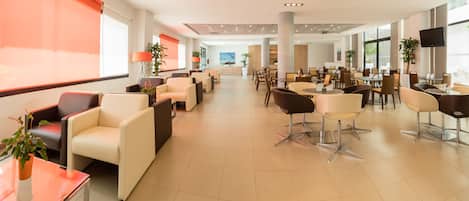 A spacious indoor area with multiple seating arrangements including sofas and chairs, large windows, and a television screen mounted on the wall.