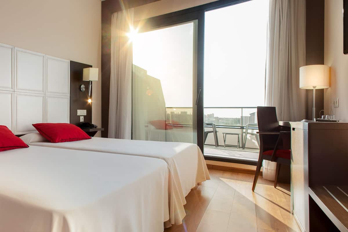 Ein Hotelzimmer mit zwei Betten, einem Schreibtisch, einem Sessel, einem Fernseher und einem Balkon mit Blick auf die Stadt.