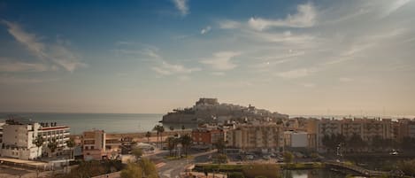 A coastal cityscape with a prominent castle-like structure on a hill, modern buildings, and a central roundabout with a small lake.