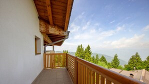 Un balcón con barandilla de madera, suelo de baldosas y vista a árboles verdes y montañas al fondo.