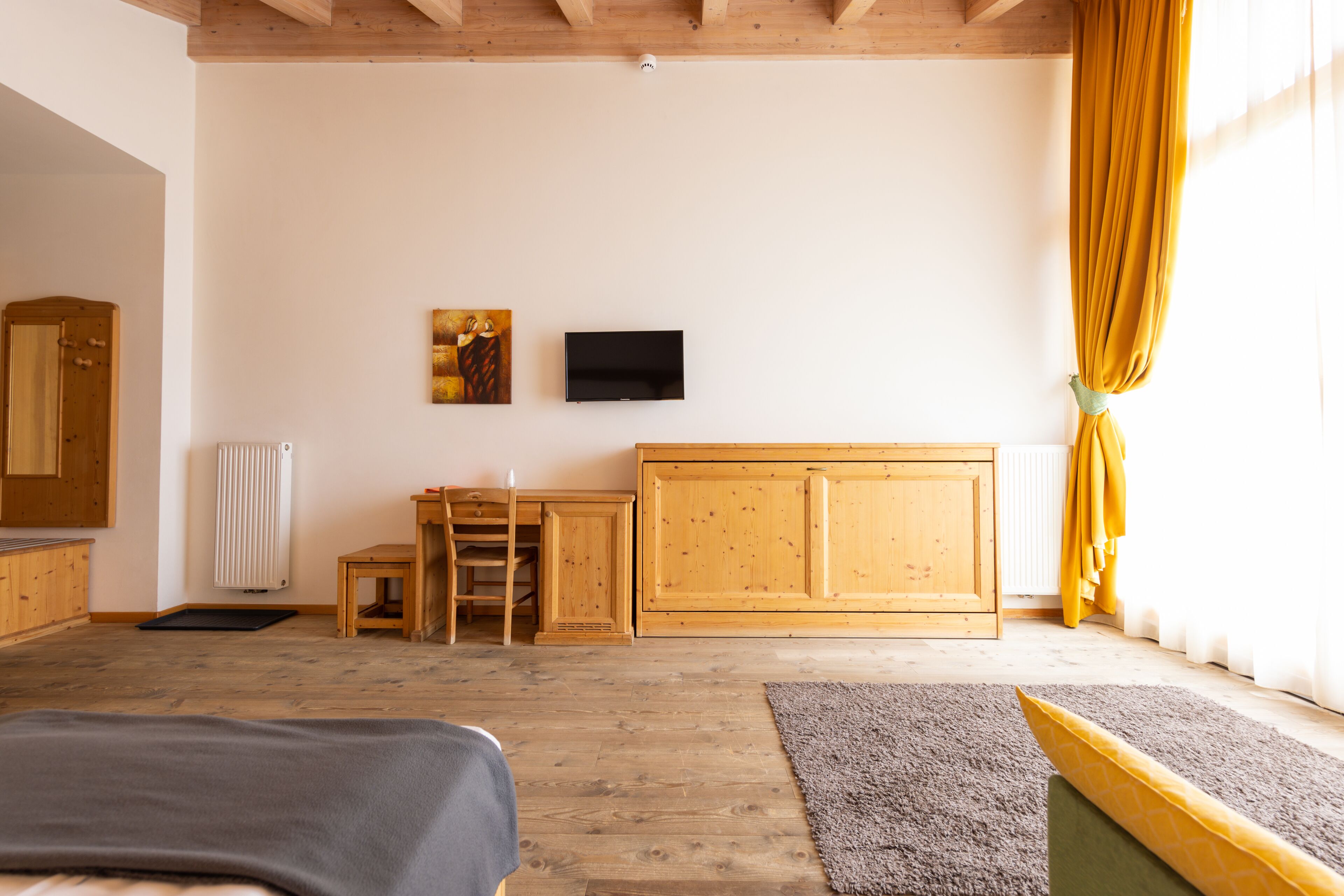 Una habitación con suelo de madera, una cama, un escritorio, una silla, un radiador, un televisor y una ventana con cortinas.