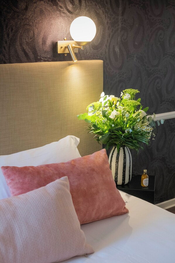 A neatly made bed with white and pink pillows, a bedside table with a vase of flowers, and a wall-mounted lamp.