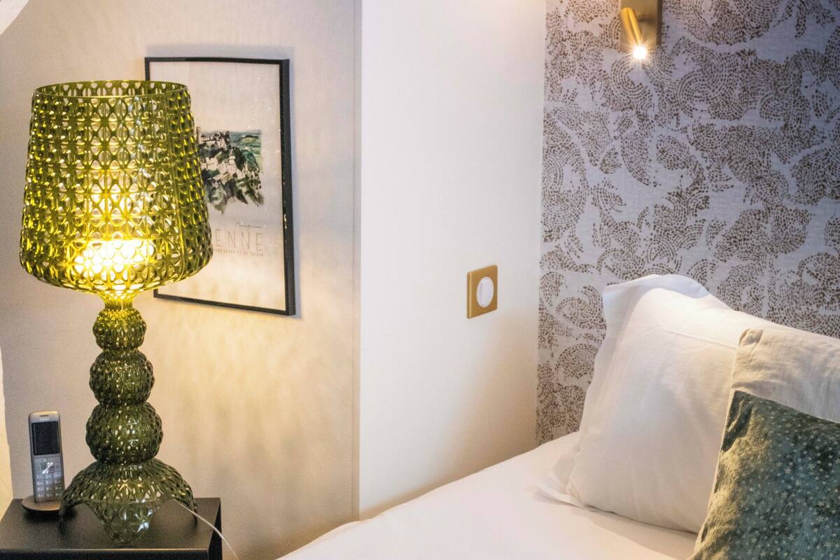 A neatly made bed with white and green pillows, a green textured lamp on a bedside table, and a wall-mounted light fixture.