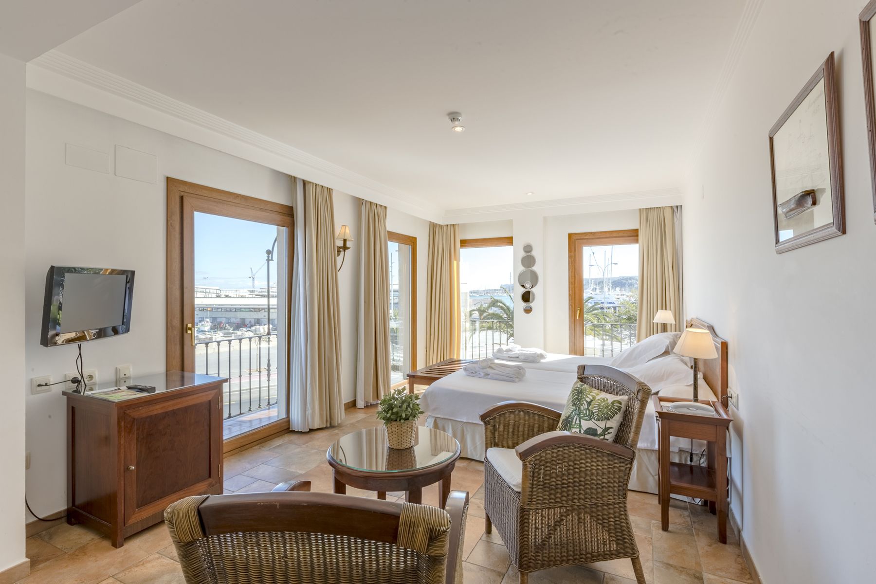 A hotel room with a bed, a TV, a desk, wicker furniture, and large windows offering a view of the city.