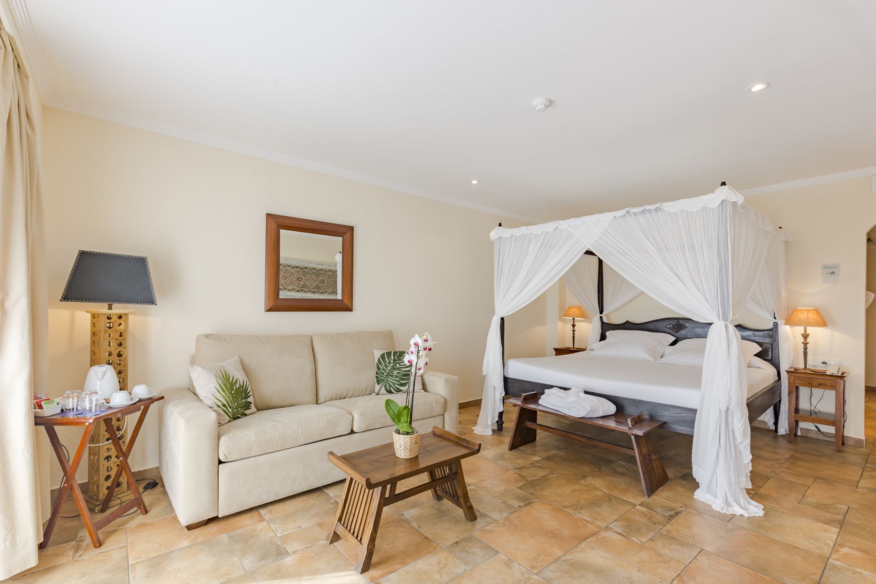 A hotel room with a four-poster bed, a sofa, a small table, and a bedside table with lamps.