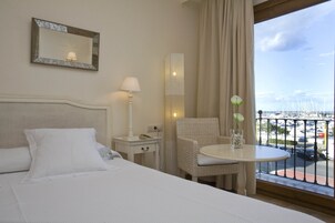 Quarto de hotel com uma cama, uma mesa pequena com uma cadeira, um abajur, um espelho e vista para uma marina através de uma varanda.
