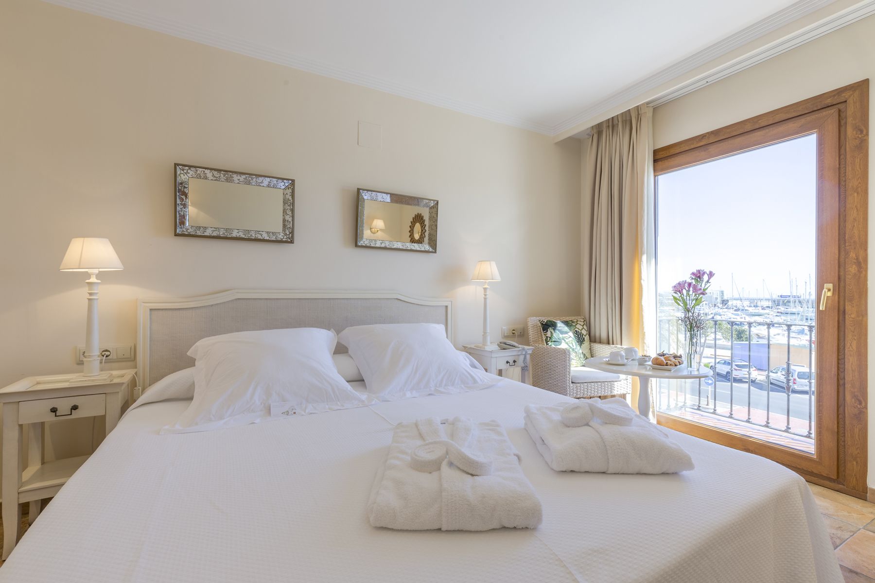 Una habitación de hotel con una cama grande, dos mesitas de noche con lámparas, una mesita pequeña con un jarrón y flores, y un balcón con vistas al puerto deportivo.