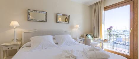 Una habitación de hotel con una cama grande, dos mesitas de noche con lámparas, una mesita pequeña con un jarrón y flores, y un balcón con vistas al puerto deportivo.