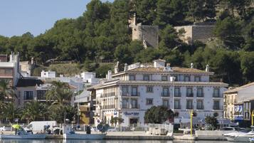 Un pueblo costero con un edificio blanco destacado con balcones, un puerto deportivo con barcos, y una colina con vegetación densa y una estructura similar a un castillo al fondo.