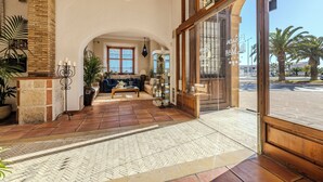 A spacious living area with a fireplace, comfortable seating, and large windows offering a view of palm trees. - La Posada del Mar Hotel (Denia)