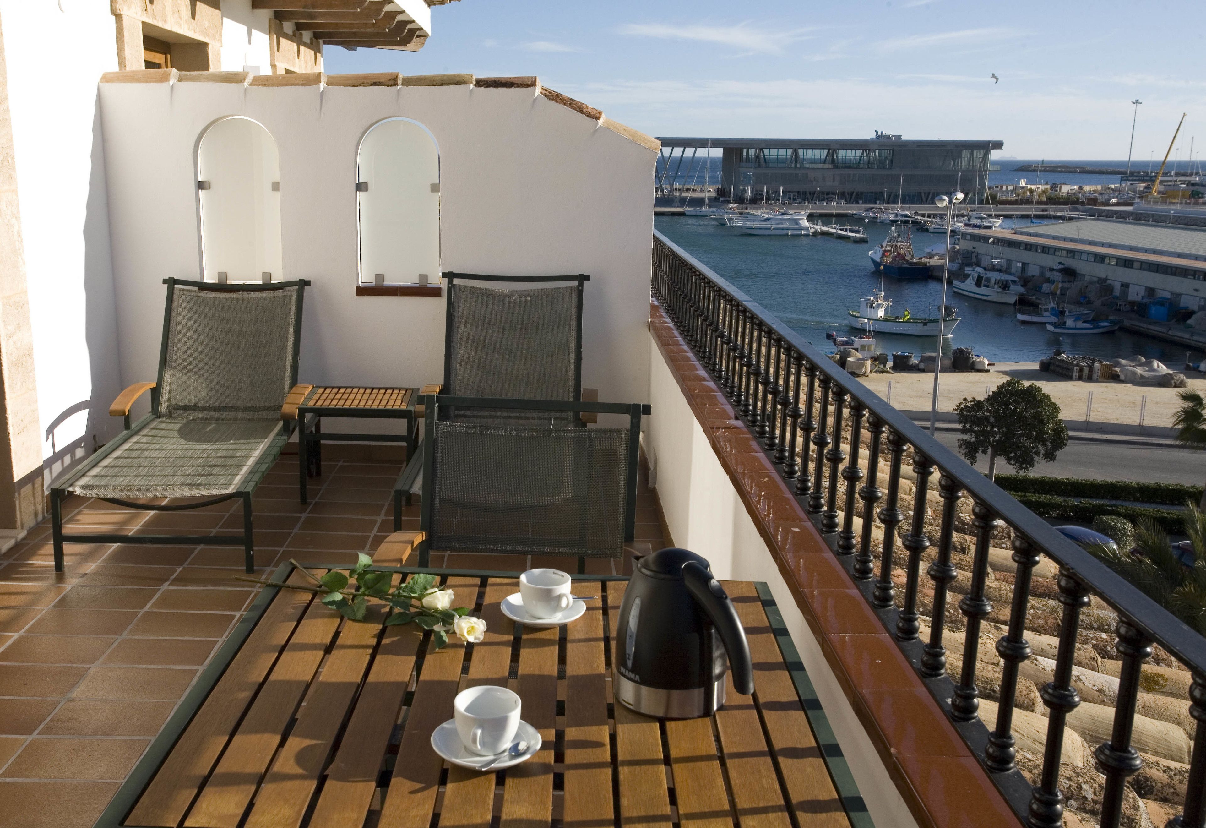 A balcony with two chairs, a table, and a kettle. The balcony overlooks a marina with boats and a bridge in the background.