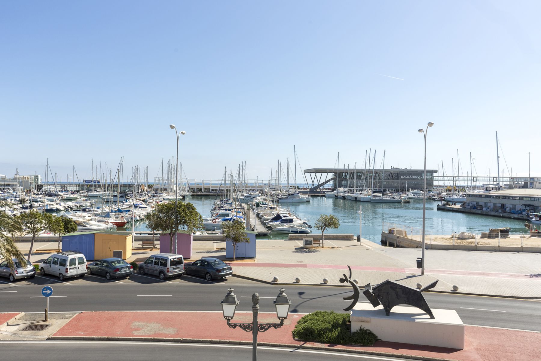 A marina with numerous boats docked, a modern building labeled 'BALEARIUM', and a street with parked cars and a pedestrian pathway.