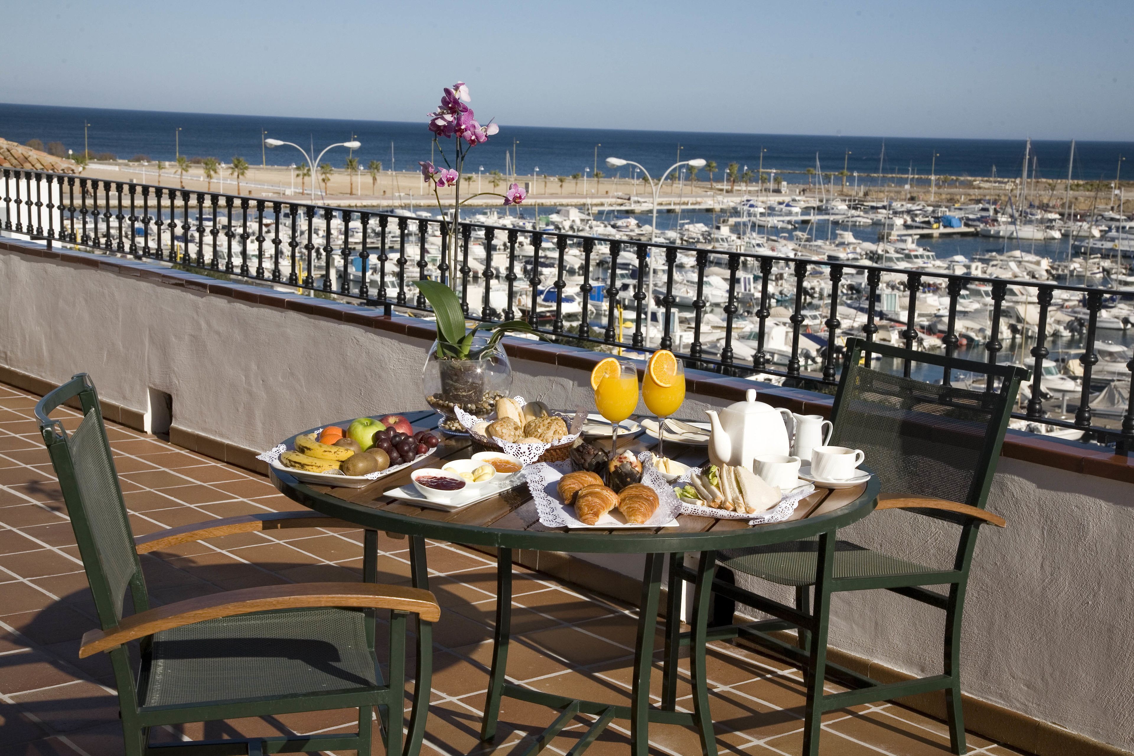 A breakfast table with a variety of food items and a vase of flowers on a balcony overlooking a marina.