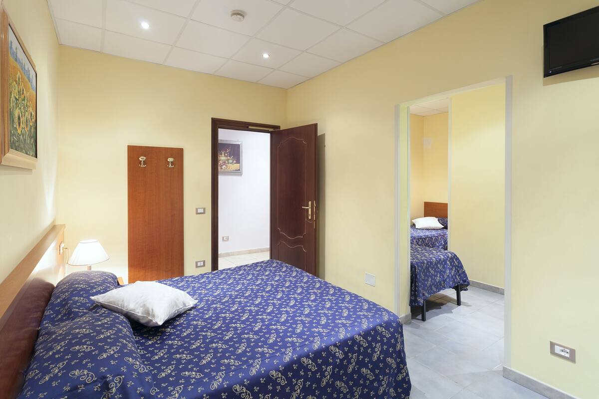 A hotel room with a bed, a nightstand, a lamp, and a door leading to another room.