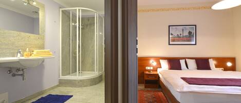 Una habitación de hotel con baño, ducha, cama y mesita de noche con lámpara.