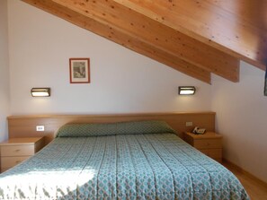 Ein Schlafzimmer mit schrägverlaufender Decke, einem Bett mit gemustertem Überwurf, zwei Nachttischen mit Lampen und einem gerahmten Bild an der Wand.