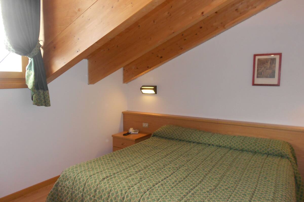 Une chambre avec un plafond en bois, un lit simple recouvert d’une couverture verte à motifs, une table de chevet avec une lampe et un tableau encadré accroché au mur.