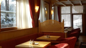 A cozy restaurant with wooden furniture, large windows with curtains, and a view of snow-covered trees. - Hotel Garni Lago Nembia (San Lorenzo Dorsino)