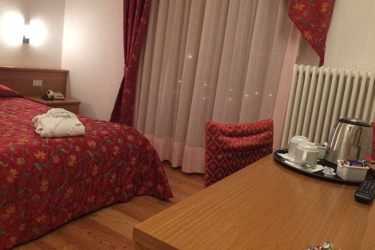 Une chambre d’hôtel avec un lit, une table de chevet avec un téléphone, une couvre-lit à motifs floraux rouges, un rideau rouge, une table de nuit en bois, un oreiller rouge, une table en bois avec un plateau contenant des articles pour le petit-déjeuner, une bouilloire et un radiateur.