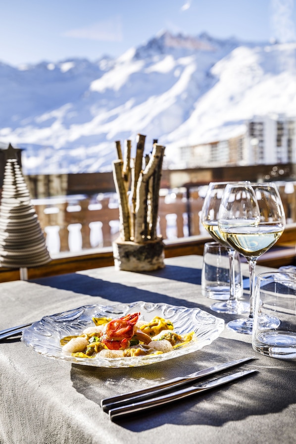 Ein Esstisch mit einem Teller Essen, einem Glas Weißwein und Blick auf schneebedeckte Berge.