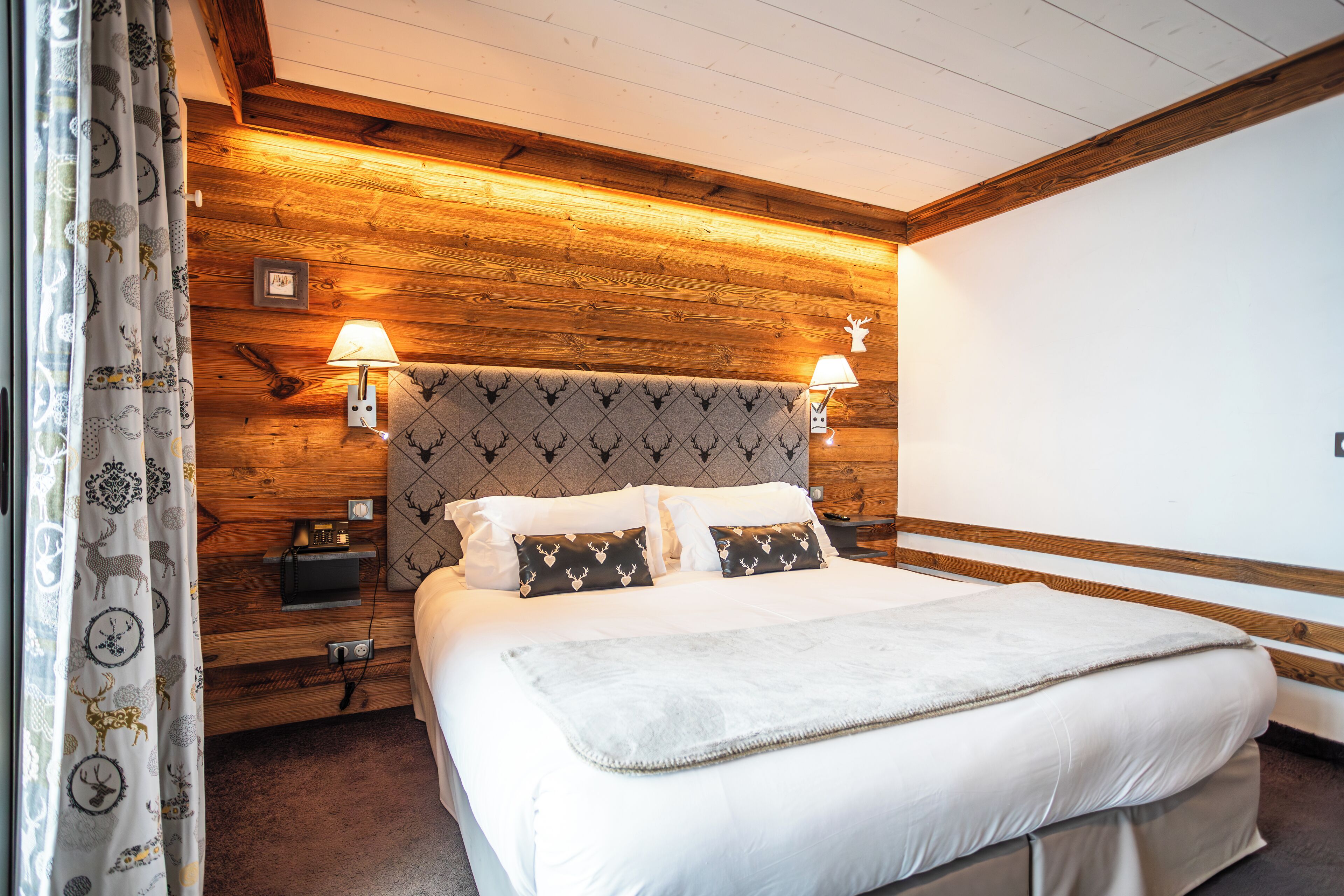 A neatly arranged bedroom with a wooden headboard, two bedside lamps, a bed with white linens, and a patterned curtain.
