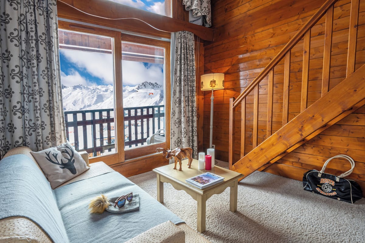 A cozy room with a wooden staircase, a sofa with a decorative pillow, a small table with a lamp, and a view of snowy mountains through large windows.