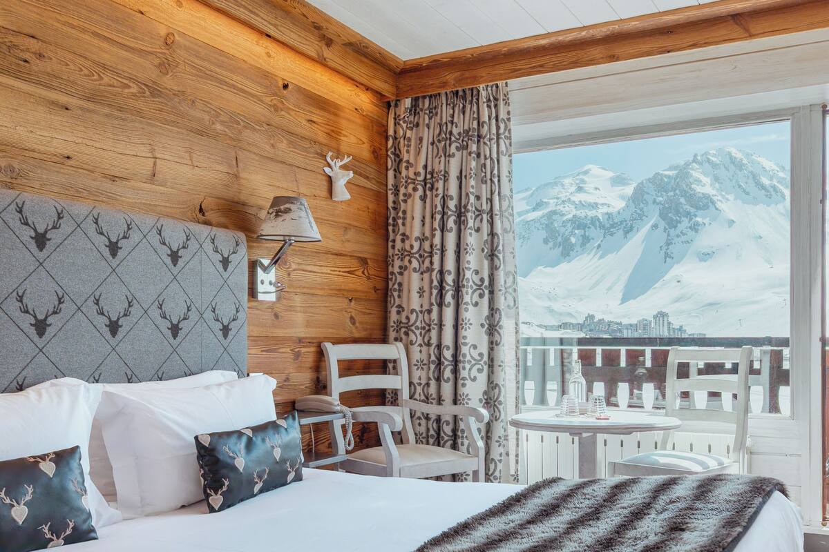 A bedroom with a large bed, a wooden headboard, a nightstand, a lamp, and a view of mountains through a balcony.