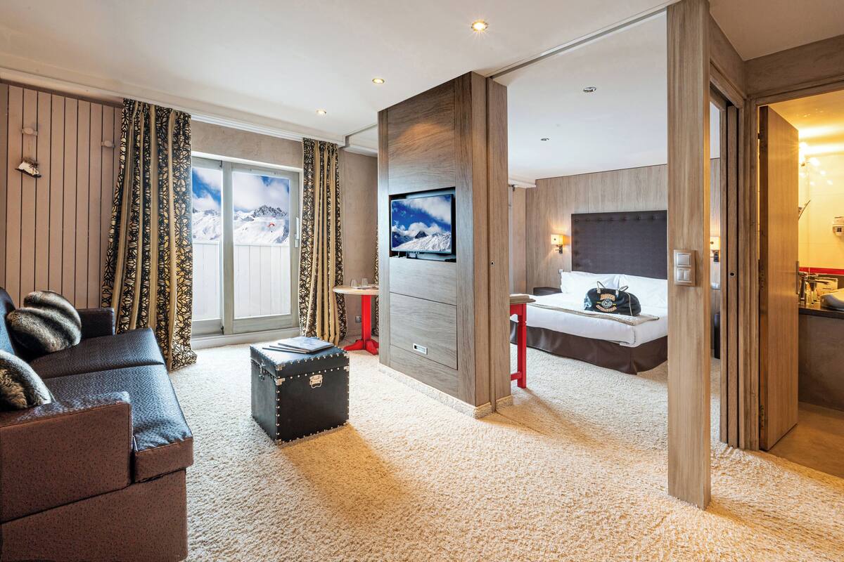 A modern hotel room with a large bed, a seating area with a sofa and ottoman, a television mounted on the wall, and a view of a snowy mountain through the window.