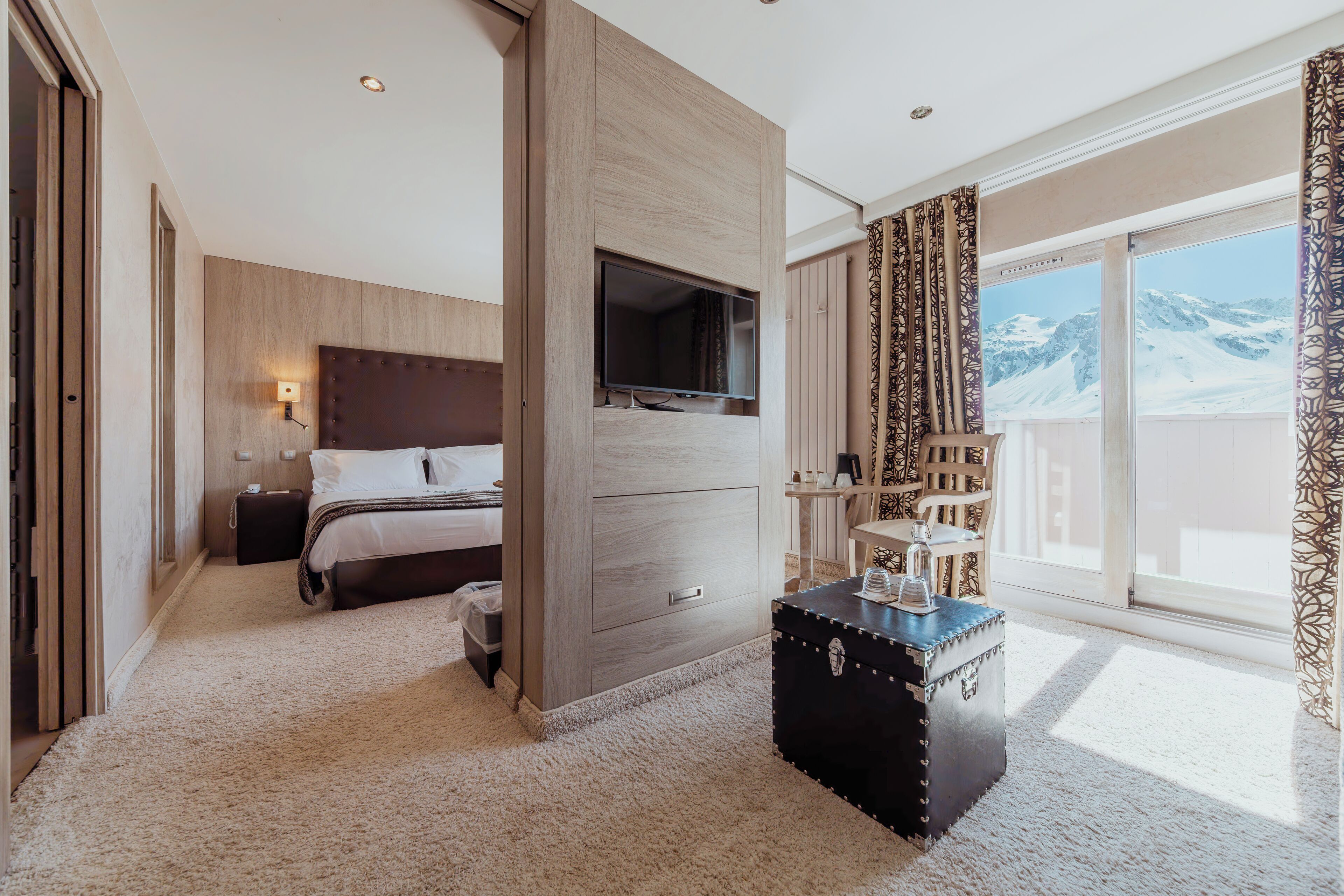 A modern hotel room with a bed, a TV mounted on a wooden panel, a small table with a decorative item, and a large window with a mountain view.