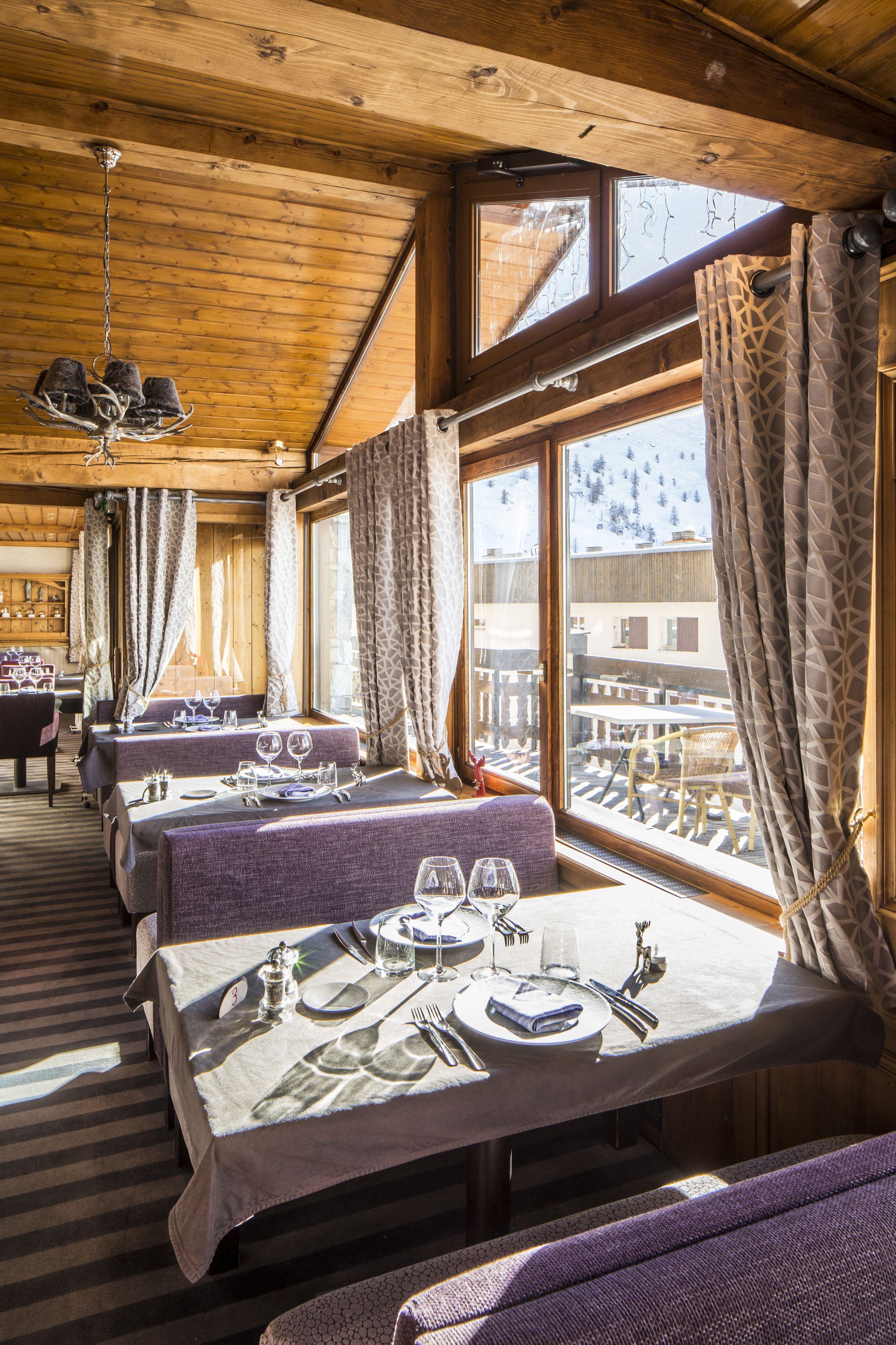A dining area with tables set for two, wooden beams on the ceiling, and large windows with curtains.