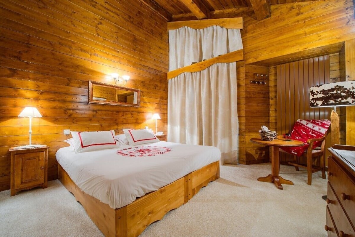 A wooden cabin bedroom with a bed, bedside lamps, a mirror, and a small table with a chair.