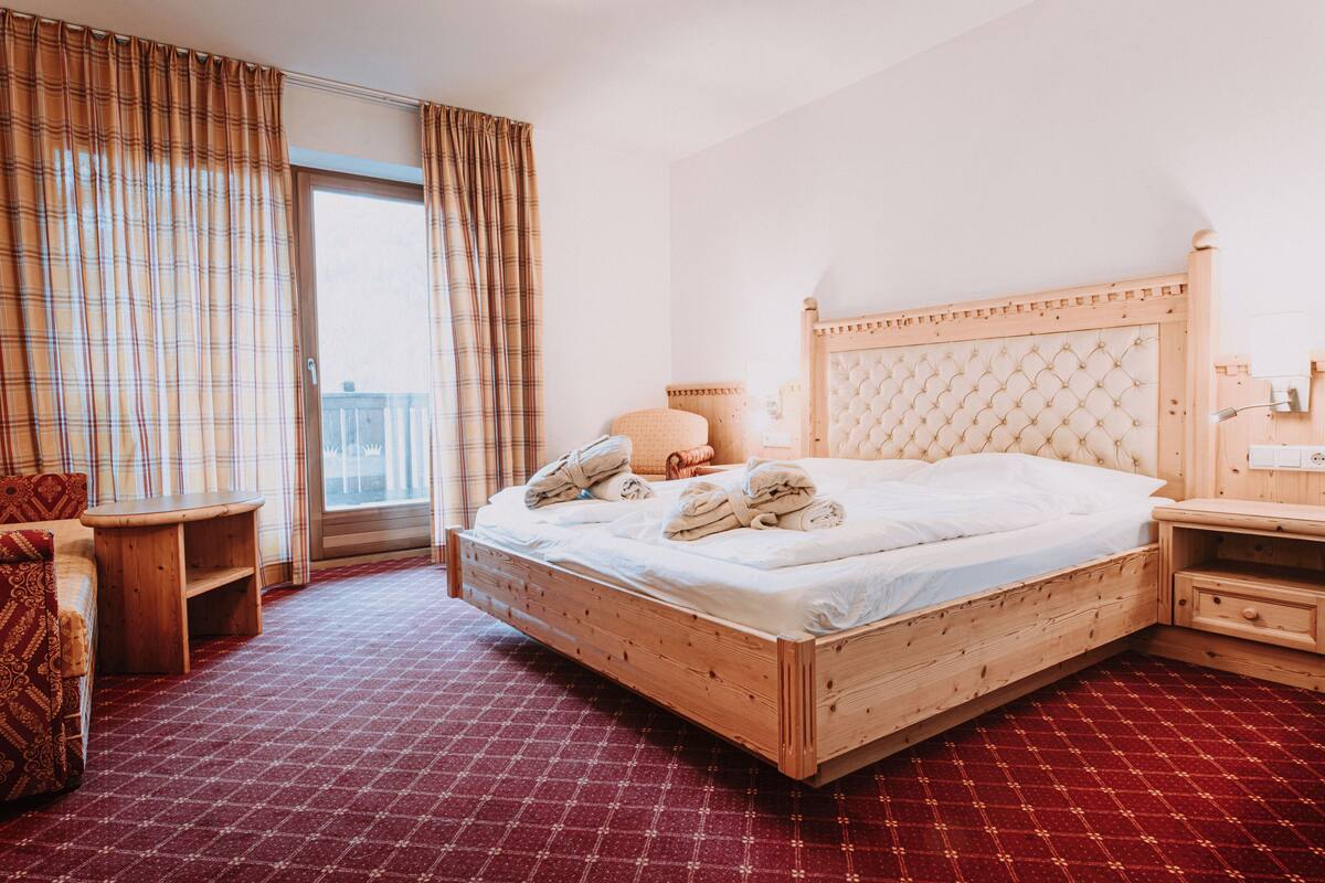 A wooden bed with a tufted headboard, a wooden nightstand, a red patterned carpet, and a window with curtains.