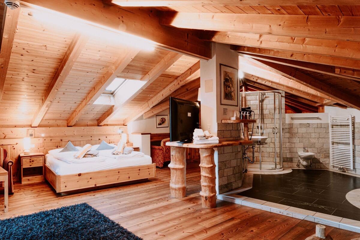 A cozy attic bedroom with a wooden ceiling, a large bed, a small table, and a bathroom with a shower and sink.