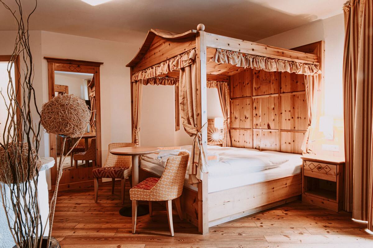 A wooden four-poster bed with a canopy, a small round table, and a chair in a room with wooden floors and walls.