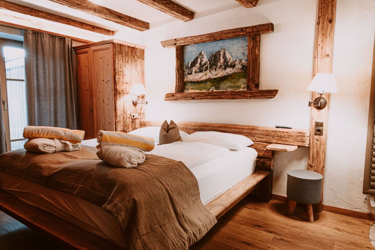 A wooden-framed bedroom with a bed, pillows, a nightstand, a lamp, and a painting of mountains.