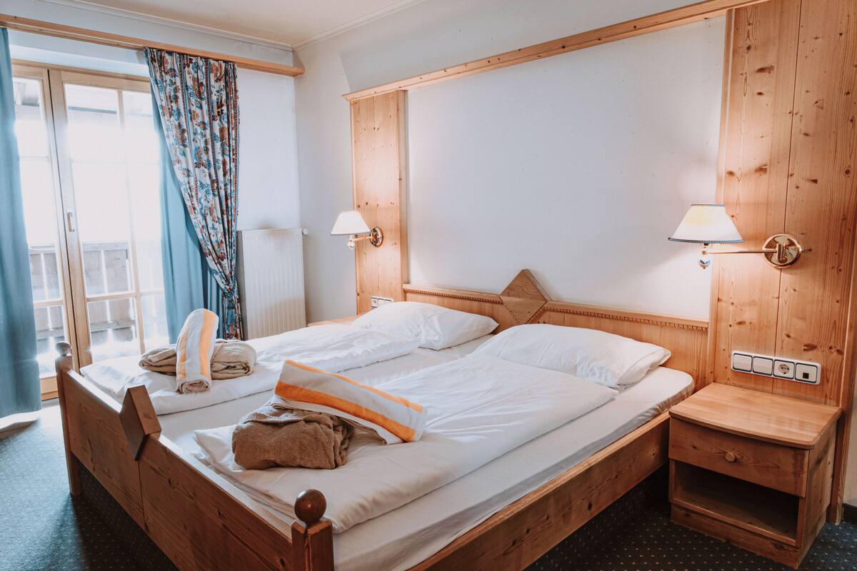 A double bed with a wooden headboard, two bedside lamps, a wooden nightstand, and a window with curtains.