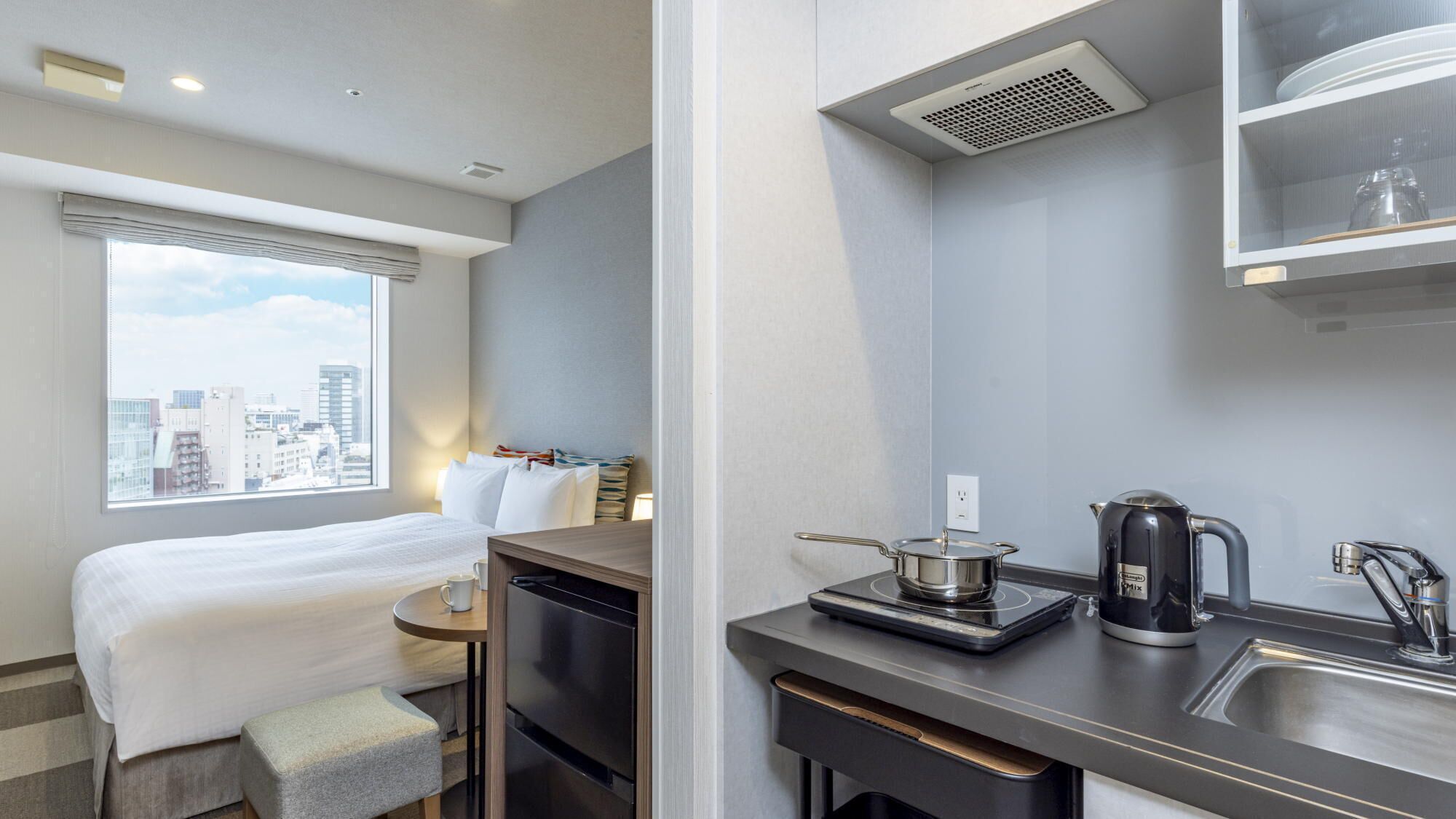 A compact hotel room with a bed, a small table, a kitchenette with a coffee maker, and a window with a city view.