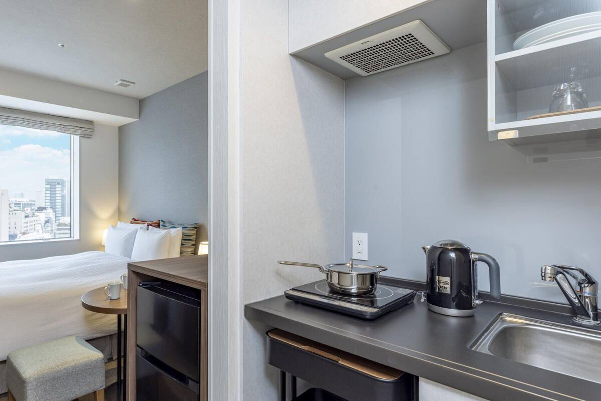 A compact hotel room with a bed, a small table, a kitchenette with a coffee maker, and a window with a city view.