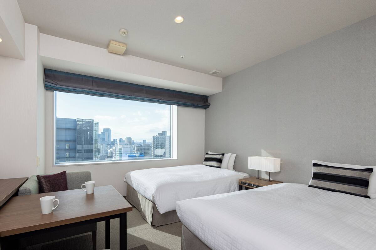 A hotel room with two beds, a desk, a chair, a window with a city view, and a lamp on the nightstand.